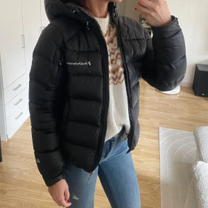 Svart dunjacka från Peak Performance - Säljer en svart dunjacka från Peak Performance. Jackan har en dragkedja framtill och en mysig huva. Perfekt för kyliga dagar och håller dig varm och bekväm.