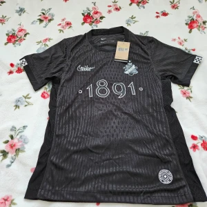 Svart fotbollströja från Nike - Säljer en svart fotbollströja från Nike med trycket '1891' på framsidan och 'SMOKINGLIRARE' på baksidan. Tröjan har korta ärmar och ett subtilt randigt mönster. Perfekt för fotbollsfans som vill ha en stilren look.