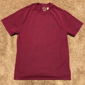 Vinröd t-shirt från Polo Ralph Lauren - Säljer en stilren vinröd t-shirt från Polo Ralph Lauren med en broderad blå logga på bröstet. T-shirten är kortärmad och tillverkad i mjuk bomull. Perfekt för en avslappnad look.