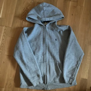 Grå hoodie från Ralph Lauren - Säljer en klassisk grå hoodie från Ralph Lauren med dragkedja och broderad logga på bröstet. Perfekt för en avslappnad stil. Den har långa ärmar och en bekväm huva.