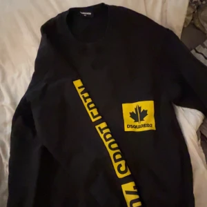 Svart tröja från Dsquared2 med gul detalj - Snygg svart tröja från Dsquared2 med en gul ficka på bröstet och text längs ärmen. Perfekt för en sportig look med en touch av stil. Tröjan har långa ärmar och en rund halsringning.