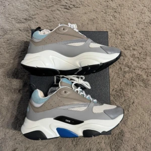 Grå och vita sneakers från Dior. Till salu för 2999 - Snygga och trendiga sneakers från Dior i grått och vitt med blå detaljer. Skorna har en chunky sula och snörning framtill. Perfekta för en stilren look.