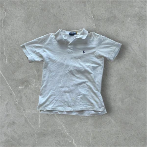 Vit pikétröja från Ralph Lauren - Säljer en klassisk vit pikétröja från Ralph Lauren med broderad logga på bröstet. Tröjan har korta ärmar och en stilren krage. Perfekt för en avslappnad men ändå stilren look. Passformen är custom slim fit. Denna tröja har två fläckar som visas på sista bilden