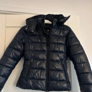 Lager 157 puffer jacka  - Snygg svart dunjacka med huva och dragkedja framtill. Jackan har en quiltad design och långa ärmar, perfekt för kyligare dagar. Praktiska fickor med dragkedja på sidorna.