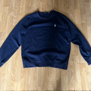 Mörkblå tröja från Polo Ralph Lauren - Säljer en stilren mörkblå tröja från Polo Ralph Lauren med deras ikoniska logga broderad på bröstet. Tröjan har långa ärmar och en klassisk rund halsringning. Perfekt för en avslappnad och snygg look.