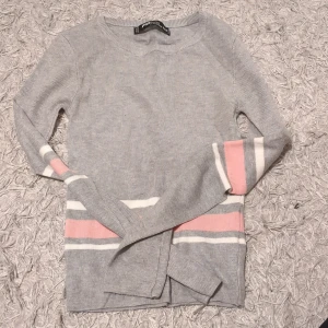 Grå ribbad tröja med rosa och vita ränder - Säljer en stilren grå ribbad tröja från Gina Tricot. Tröjan har långa ärmar och dekorativa rosa och vita ränder vid ärmslut och nederkant. Perfekt för en avslappnad look.