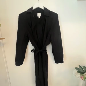 Svart kappa från H&M storlek xs - Snygg svart kappa från H&M med långa ärmar och knytband i midjan. Perfekt för en stilren look. Kappan är i storlek xs men är stor i storlek 