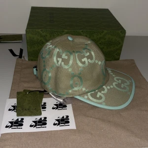Beige och mintgrön keps från Gucci Jumbo - Snygg keps från Gucci i beige med mintgröna detaljer och det ikoniska GG-mönstret. Kepsen har en böjd skärm och justerbar rem baktill för perfekt passform. Perfekt för att ge din outfit en lyxig touch.