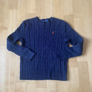 Mörkblå stickad tröja från Ralph Lauren - Säljer en snygg mörkblå stickad tröja från Ralph Lauren med kabelstickat mönster. Tröjan har långa ärmar och en broderad logotyp i orange på bröstet. Perfekt för kyliga dagar!