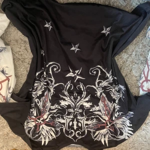 Svart tröja med mönster - Säljer en svart tröja med ett detaljerat vitt och rött mönster. Tröjan har långa ärmar och ett unikt tryck med stjärnor och blommor. Perfekt för dig som vill ha något annorlunda i garderoben!