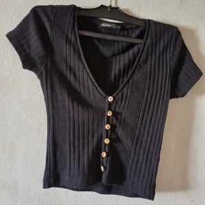 Svart ribbad topp från Pull&Bear - Säljer en svart ribbad topp från Pull&Bear med korta ärmar och v-ringning. Toppen har knappar framtill i olika färger som ger en unik touch. Perfekt för en stilren look.