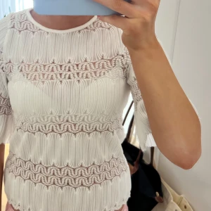 Vit blus med spetsdetaljer - Säljer en elegant vit blus med vackra spetsdetaljer och korta ärmar. Blusen har ett unikt mönster med vågformade strukturer och transparenta partier som ger en stilren look. Perfekt för att lyfta vilken outfit som helst. Knappt använd!