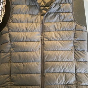 Grå dunväst från Uniqlo - Säljer en stilren grå dunväst från Uniqlo. Västen är ultralätt och har en dragkedja framtill. Perfekt för lager-på-lager under kyliga dagar. Den är tillverkad med vattenavvisande material för extra skydd mot väder och vind.