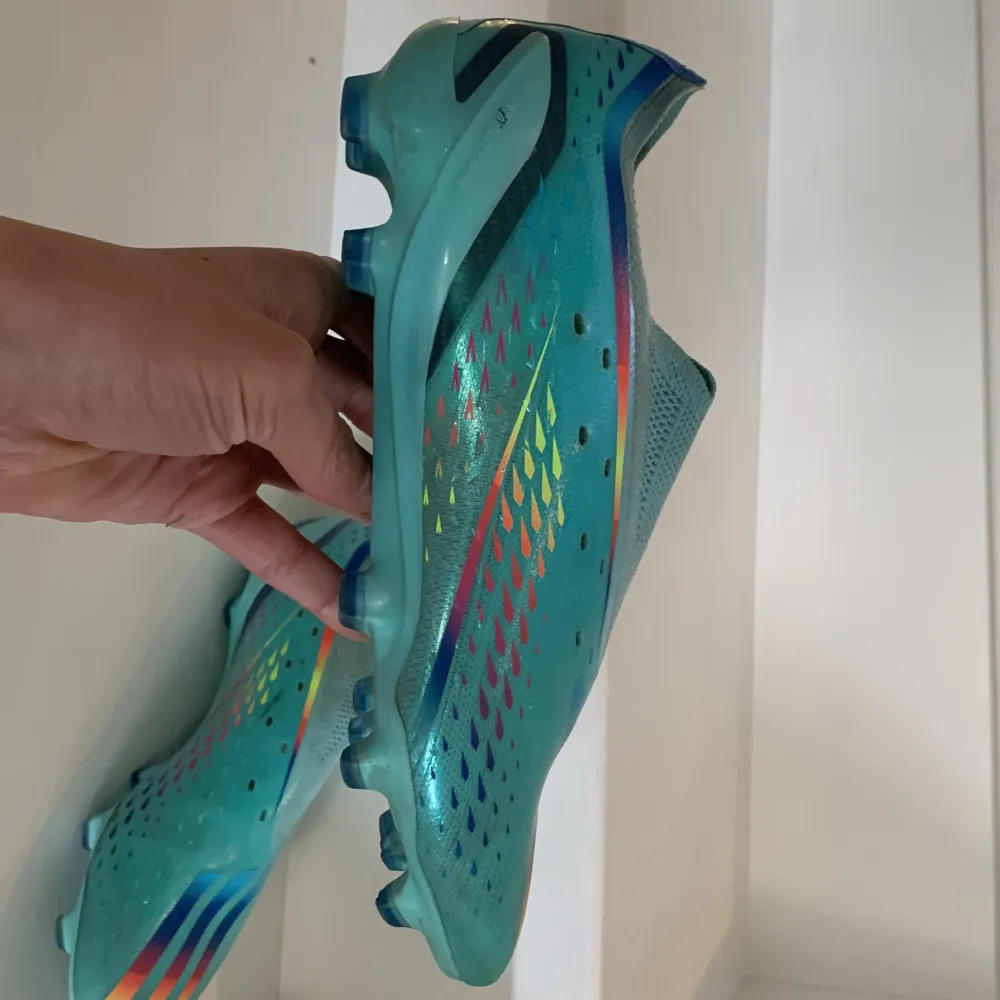 Snygga turkosa fotbollsskor med färgglada regnbågsdetaljer och snörning. Skorna har en rund tå och är designade för att ge bra grepp på planen. Perfekta för fotbollsspelare som vill sticka ut. . Kengät.