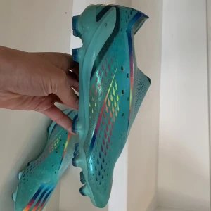 Adidas X speedportal  - Snygga turkosa fotbollsskor med färgglada regnbågsdetaljer och snörning. Skorna har en rund tå och är designade för att ge bra grepp på planen. Perfekta för fotbollsspelare som vill sticka ut. 