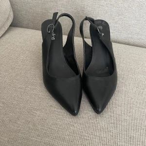 Svarta pumps från Ellos stl 37 - Snygga svarta pumps från Ellos med spetsig tå och smal klack. Perfekta för en elegant look. De har en stilren design med en justerbar rem runt hälen.