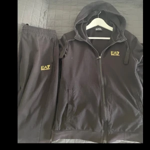 EA7 tracksuit  - Säljer en stilren svart hoodie från EA7 med dragkedja och huva. Tröjan har en gul logotyp på bröstet och är perfekt för en avslappnad look. Den har långa ärmar och praktiska fickor framtill. Byxa följer även med ea7
