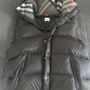 Svart dunväst med mönstrad krage - Snygg svart dunväst från Burberry med en klassisk rutig krage i grått, vitt och rött. Västen har dragkedja och knappar framtill samt praktiska fickor med dragkedja. Perfekt för kyliga dagar!