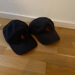 Svart keps från Polo Ralph Lauren - Säljer två svarta kepsar från Polo Ralph Lauren med röd broderad logga fram och texten 'POLO' bak. Kepsarna är tillverkade i 100% bomull och har en klassisk design med justerbar rem.