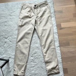 Beige linnebyxor från John Henric - Snygga beige linnebyxor från John Henric med knappar och snörning i midjan. Perfekta för en stilren och avslappnad look. Byxorna har en normal passform och är tillverkade i ett lätt och luftigt material.