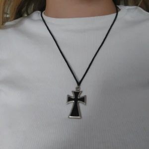 Svart korshalsband - Snyggt halsband med ett svart korshänge. Hänget har en silverkant och hänger på ett svart snöre. 