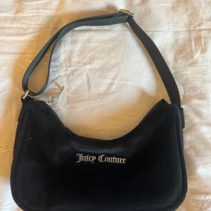 Svart axelväska från Juicy Couture - Snygg svart axelväska från Juicy Couture i mjukt material. Väskan har en dragkedja upptill och ett stilrent märke på framsidan. Perfekt för att bära dina nödvändigheter med stil.