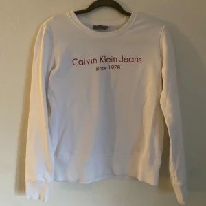 Vit tröja från Calvin Klein Jeans - Säljer en stilren vit tröja från Calvin Klein Jeans med röd text på bröstet. Tröjan har långa ärmar och en klassisk rund halsringning. 