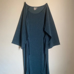 Blå klänning från ICHI - Snygg blå klänning från ICHI med glittriga detaljer. Den har en lös passform och trekvartsärmar, perfekt för en stilren look. Klänningen är tillverkad i ett bekvämt material som ger en elegant känsla. Den har även ett snöre man kan knyta runt midjan.