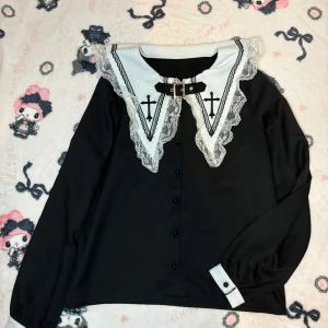 Svart blus med spetskrage och korsdetaljer - Snygg svart blus med en stor vit spetskrage dekorerad med kors och en spänndetalj. Blusen har långa ärmar och knappar framtill. Perfekt för jirai kei och andra jfashion stilar. Från ma*rs