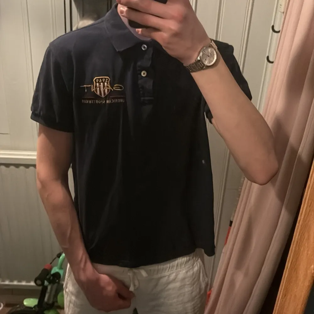 Säljer en stilren mörkblå pikétröja från GANT med korta ärmar och klassisk krage. Tröjan har en broderad logotyp i guld på bröstet. Perfekt för en avslappnad men ändå snygg look.. T-paidat.