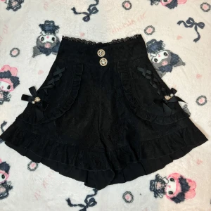 Svarta spets shorts från Rojita - Snygga svarta shorts i spets från Rojita med dekorativa knappar och rosetter. Shortsen har volangdetaljer och en hög midja med spetskant. Perfekta för ryousangata och jirai kei!