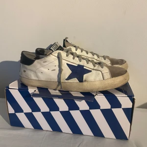 Golden Goose sneakers i Vitt med blå stjärna   - Säljer nu dessa riktigt sköna Golden Goose skorna i en ovanlig färg (nypris ca 6000kr). Skorna har synliga hål vid hälen men det är inget en som skomakare inte kan fixa.                                 Original box medföljer, självklart äkta.                           Hör gärna av her vid övriga frågor ;).                           Priset är ej satt i sten!