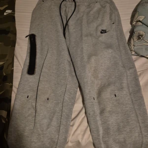 Grå och kamouflage mjukisbyxor från Nike - Säljer två par mjukisbyxor från Nike. Ett par är gråa med svarta detaljer och ett par är i kamouflagemönster. Båda har elastisk midja med snörning och Nike-logga på sidan.