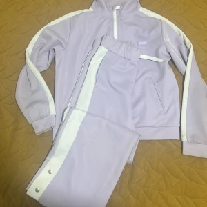 Ljuslila träningsset  - Snyggt ljuslila träningsset  med vita detaljer. Jackan har dragkedja och långa ärmar, medan byxorna har knappar längs sidorna. Perfekt för en sportig look!