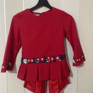 Röd blus med en prickig kjol - Charmig klänning i rött och blått med vita prickar. Överdelen är röd med blommiga broderier och volanger, medan kjolen är blå med vita prickar. Långärmad design med dragkedja bak och knytband i midjan.