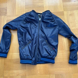 Blå bomberjacka med dragkedja - Snygg blå bomberjacka från GAP med dragkedja och ribbade muddar i blått och svart. Jackan har två fickor med dragkedja framtill och en klassisk bomberstil. Perfekt för en cool och avslappnad look.