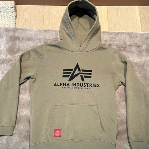 Alpha Industries hoodie grön - Säljer en olivgrön hoodie från Alpha Industries med stor logga på framsidan. Tröjan har en klassisk känguruficka och ribbade muddar vid ärmslut och nederkant. Perfekt för en avslappnad stil.