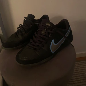 Svarta Nike sneakers med blå detalj - Snygga svarta Nike sneakers med en iögonfallande blå swoosh på sidan. Skorna har snörning och en rund tå, vilket ger en klassisk och bekväm passform. Perfekta för en sportig look.