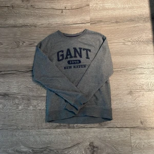 Grå långärmad tröja från GANT - Säljer en stilren grå långärmad tröja från GANT med tryck 'GANT 1949 NEW HAVEN' i mörkblått på framsidan. Perfekt för en avslappnad stil. Tröjan har en rund halsringning och är tillverkad i ett mjukt material.