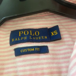 Rosa och vit randig skjorta från Ralph Lauren - Säljer en stilren rosa och vit randig skjorta från Ralph Lauren. Skjortan har en klassisk krage och långa ärmar med knappar vid ärmsluten. På sista bilden ser du en fläck, det finns en sån fläck på två ställen, den är väldigt liten och syns knappt!! 