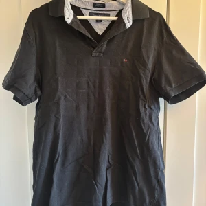 Svart pikétröja från Tommy Hilfiger - Säljer en stilren svart pikétröja från Tommy Hilfiger med klassisk krage och korta ärmar. Tröjan har en liten broderad logotyp på bröstet och kontrasterande detaljer på insidan av kragen. 