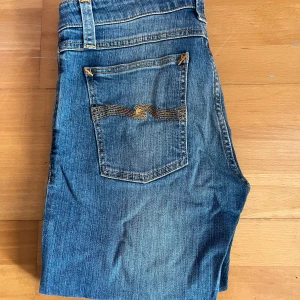 Blå jeans från Nudie Jeans - Säljer ett par klassiska blå jeans från Nudie Jeans med snygga orangea sömmar och femficksdesign. Jeansen har en slim passform och är tillverkade i slitstarkt denim. Perfekta för en avslappnad stil.