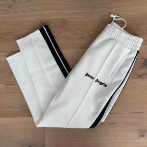 Vita byxor från Palm Angels - Snygga vita byxor från Palm Angels med svarta ränder längs sidorna. Byxorna har en elastisk midja med snörning och en stilren design med logotypen tryckt på framsidan. Perfekta för en avslappnad och trendig look.