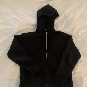 Svart hoodie ifrån ACNE STUDIOS med dragkedja - Säljer en klassisk svart hoodie med dragkedja och huva. Perfekt för en avslappnad stil. Den har långa ärmar och en praktisk ficka framtill. Passar till alla tillfällen!