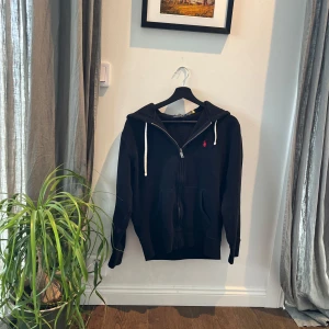 Ralph Lauren hoodie - Säljer en stilren svart hoodie från Polo Ralph Lauren med dragkedja och en liten röd logotyp på bröstet. Perfekt för en avslappnad look. Den har långa ärmar och en justerbar huva med vita snören. Storlek S 9/10 skick inga skador alls. Väldigt bra passform och sjukt snygg. Kom med pris förslag