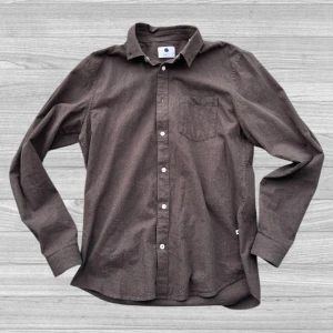 NN07 skjorta - Säljer en stilren brun skjorta från NN07 i storlek M. Skjortan har långa ärmar och en klassisk krage med knappar framtill. Perfekt för en avslappnad men ändå uppklädd look.