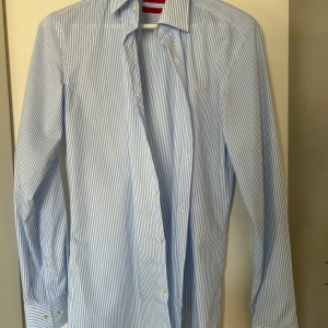 Blå och vit randig skjorta från Hugo Boss - Snygg blå och vit randig skjorta från Hugo Boss i slim fit. Skjortan har långa ärmar och knappar framtill. Perfekt för en stilren look.