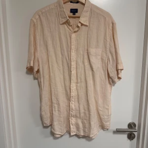 Beige linneskjorta från GANT - Säljer en stilren beige kortärmad linneskjorta från GANT. Skjortan har en klassisk krage och knappar framtill samt en bröstficka med broderad logotyp. Perfekt för varma dagar.