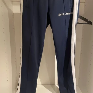Mörkblå träningsbyxor från Palm Angels - Snygga mörkblå träningsbyxor från Palm Angels med vita sidoremsor och elastisk midja. Perfekta för en sportig look eller avslappnad stil. Byxorna har en klassisk passform och är tillverkade i ett bekvämt material.