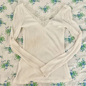 Vit ribbad långärmad topp med spetsdetalj - Säljer en stilren vit ribbad topp med vacker spetsdetalj vid halsringningen. Toppen har långa ärmar och en elegant v-ringning. Perfekt för en chic och bekväm look.